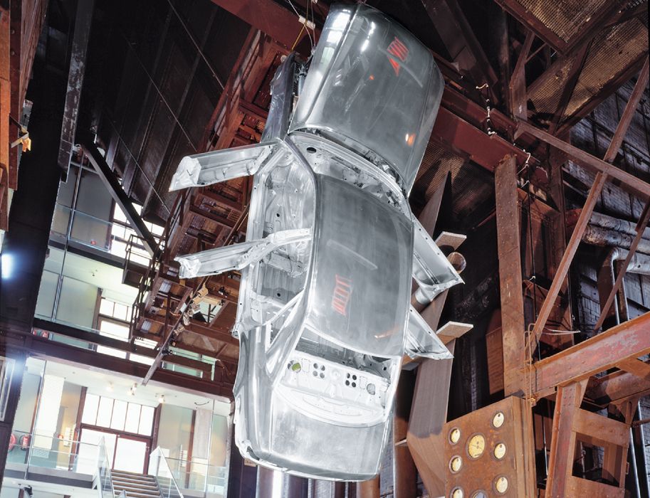 The bare aluminum body of an Audi A8 hangs from the ceiling between steel beams in an exhibition hall